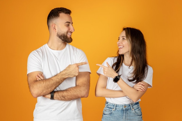 Młody mężczyzna i młoda kobieta, obaj w białych koszulkach i uśmiechnięci, zabawnie wskazują na siebie