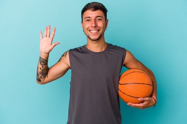 Młody mężczyzna kaukaski gry w koszykówkę na białym tle na niebieskim tle uśmiechający się wesoły pokazując numer pięć palcami.