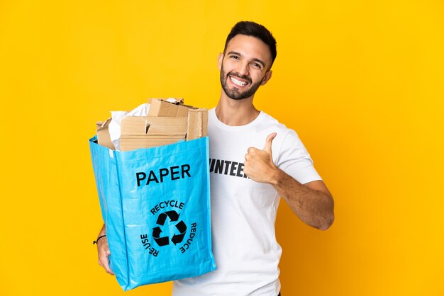 Młody mężczyzna kaukaski trzymając torbę recyklingu pełnego papieru do recyklingu na białym tle, dając kciuk w górę gestu