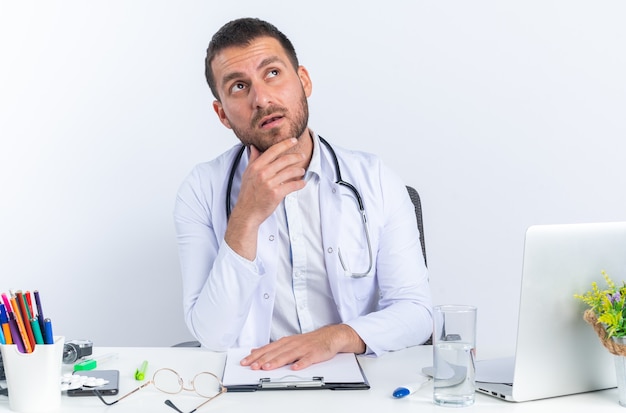 Młody mężczyzna lekarz w białym fartuchu i stetoskopem patrząc z ręką na brodzie z zamyślonym wyrazem twarzy siedzącej przy stole z laptopem na białym tle