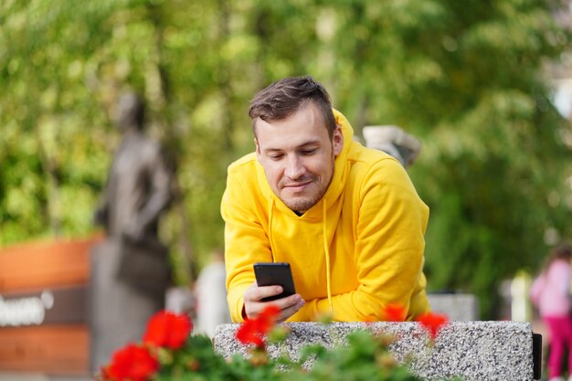 Młody mężczyzna leżący na brzuchu na ławce z podniesionymi nogami i przeglądający telefon komórkowy Mężczyzna korzystający ze smartfona korzysta z Internetu na dziedzińcu budynku mieszkalnego