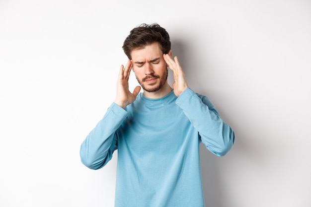 Młody mężczyzna mający ból głowy lub kaca, dotykający skroni głowy i marszczący brwi, mdłości z powodu migreny, stojący na białym tle