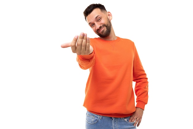 Młody mężczyzna na białym tle w pomarańczowym swetrze wyciąga rękę do przodu kiwa ku niemu