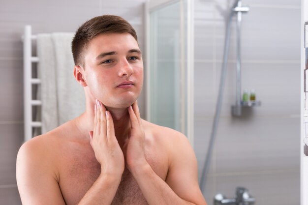Młody mężczyzna nałożył na twarz balsam po goleniu dwiema rękami stojąc przed lustrem w nowoczesnej, wyłożonej kafelkami łazience w domu