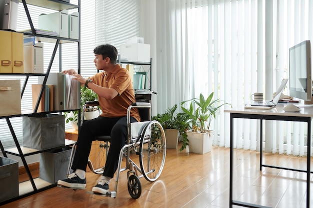 Młody mężczyzna niepełnosprawny poruszający się po biurze domowym na wózku inwalidzkim, szukając dokumentów