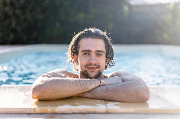Młody mężczyzna oparty na basenie z rękami skrzyżowanymi podczas letnich wakacji Kaukaski facet sam w basenie Koncepcja pielęgnacji skóry i ochrony przeciwsłonecznej