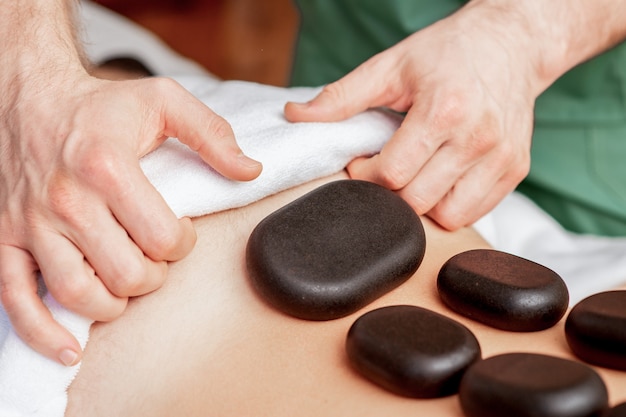 Młody mężczyzna otrzymuje masaż kamieniami na plecach, podczas gdy ręce masażysty kładzie kamienie na jej plecach.