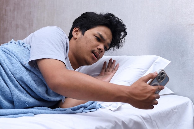 Młody mężczyzna patrzący na budzik na smartfonie, leżąc na łóżku w sypialni ze zszokowanym byłym