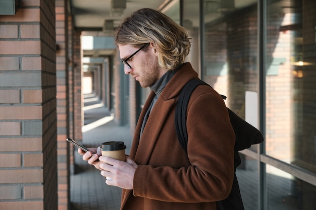 Młody Mężczyzna Pisze Wiadomość Przez Telefon I Pije Kawę Stojąc Na Zewnątrz