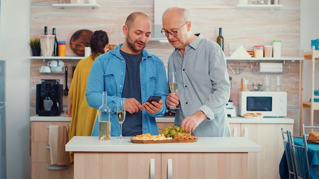 Młody mężczyzna pokazuje ojcu, jak korzystać z nowoczesnego smartfona, siedząc w kuchni i popijając kieliszek białego wina przed rodzinnym obiadem. Nowy styl życia, korzystanie z technologii, podczas gdy kobiety przygotowują uzdrowienie