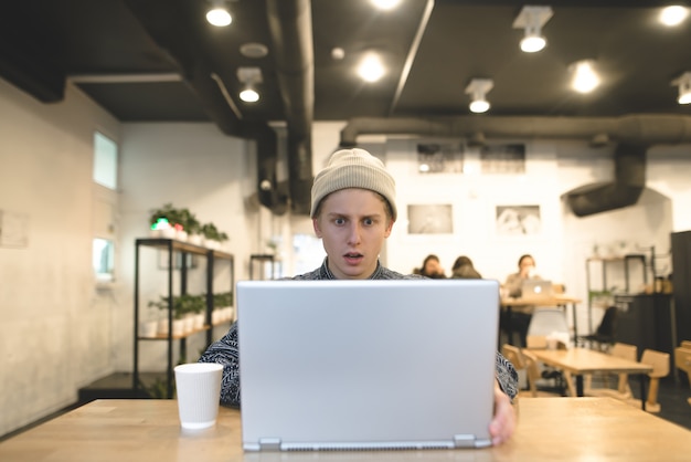 Zdjęcie młody mężczyzna pracuje w kawiarni i wygląda na zaskoczonego ekranem laptopa. uczeń korzysta z internetu w przytulnej kawiarni.