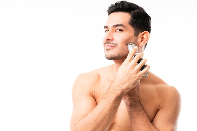 Młody mężczyzna przycinający brodę elektryczną maszynką do golenia w studio