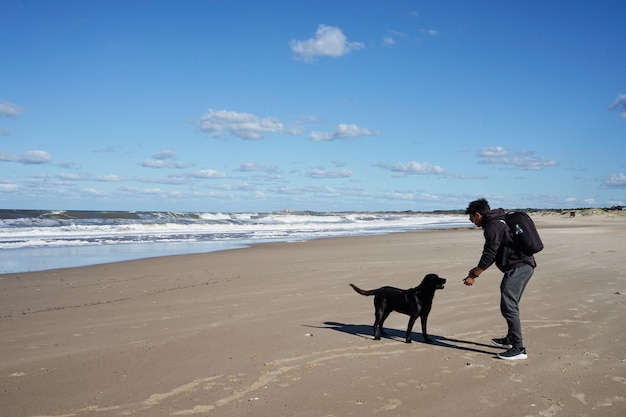 Młody mężczyzna rasy kaukaskiej grając na plaży z czarnym psem. Na piasku pod błękitnym niebem z chmurami.