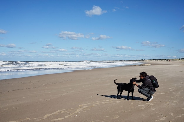 Młody Mężczyzna Rasy Kaukaskiej Grając Na Plaży Z Czarnym Psem. Na Piasku Pod Błękitnym Niebem Z Chmurami.