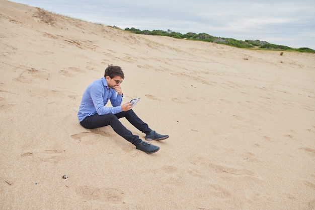 Młody mężczyzna rasy kaukaskiej myślenia podczas czytania swojego notebooka, siedząc na piasku na plaży. Ma na sobie garnitur.