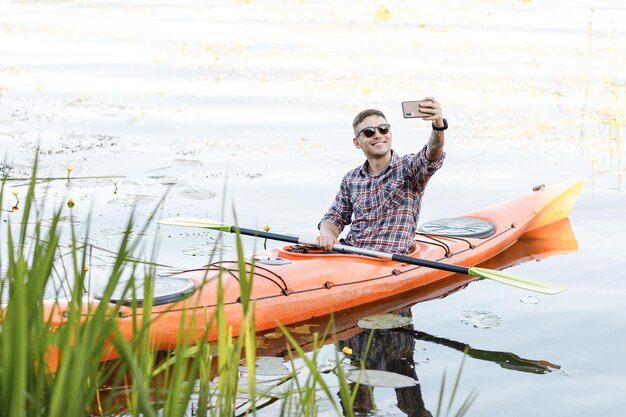 Młody mężczyzna rasy kaukaskiej siedzi w kajaku i robi selfie na telefonie komórkowym Koncepcja rozrywki wodnej