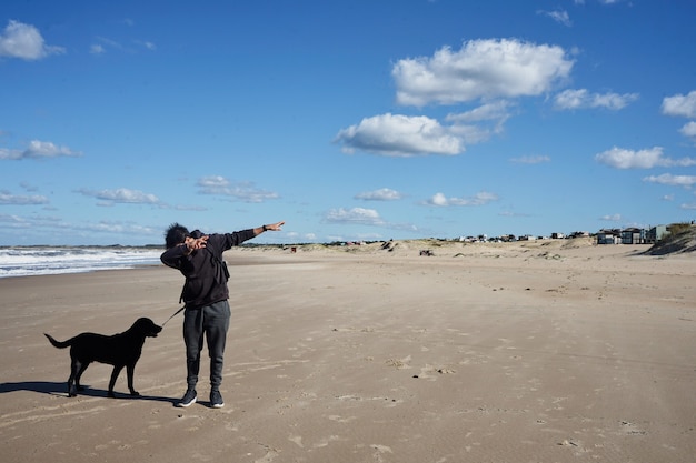 Młody mężczyzna rasy kaukaskiej z czarnym psem na plaży pod błękitnym pochmurnym niebem.