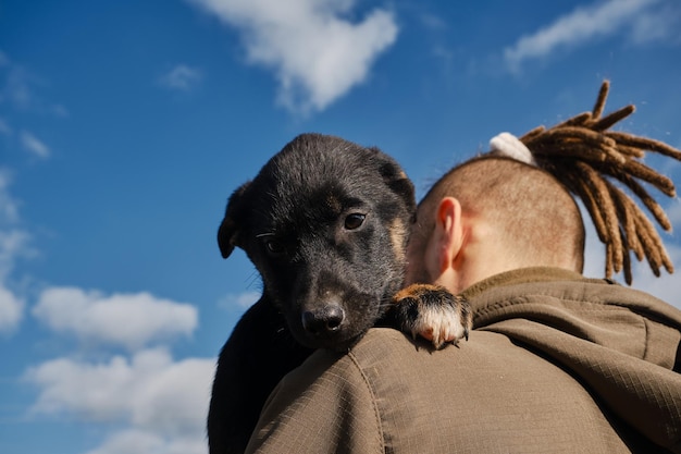 Młody mężczyzna rasy kaukaskiej z dredami przytula czarnego szczeniaka husky alaskańskiego na zewnątrz w ciepły dzień