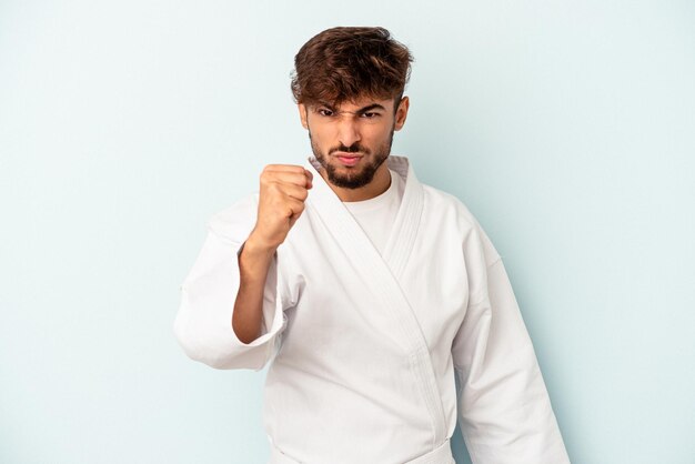 Młody mężczyzna rasy mieszanej robi karate na białym tle na niebieskim tle pokazując pięść do kamery, agresywny wyraz twarzy.