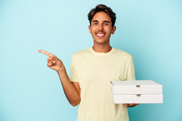 Młody mężczyzna rasy mieszanej trzymający pizze na białym tle na niebieskim tle, uśmiechając się i wskazując na bok, pokazując coś w pustej przestrzeni.
