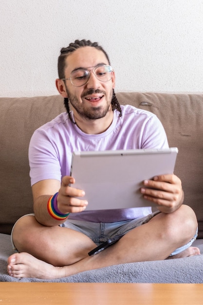 Młody mężczyzna siedzący na kanapie używający swojego tabletu Widok z przodu mężczyzny siedzącego w salonie i używającego tabletu Uśmiechnięty mężczyzna dotykający cyfrowego tabletu siedzącego w pokoju