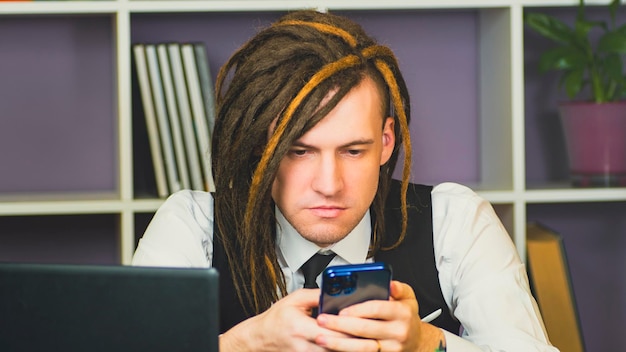 Młody mężczyzna siedzący przy laptopie przy użyciu smartfona w domowym biurze Nowoczesny freelancer przeglądający telefon komórkowy w miejscu pracy w domu