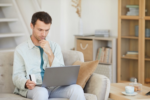 Młody mężczyzna siedzi na kanapie z laptopem ma zamiar zapłacić w domu kartą kredytową w trybie online