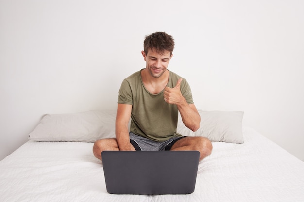 Młody mężczyzna siedzi na łóżku na rozmowie wideo ze swoim laptopem, kciuki do góry pozytywne jak znak i ziewanie. Nosi zieloną koszulę. Łóżko i biały pokój.