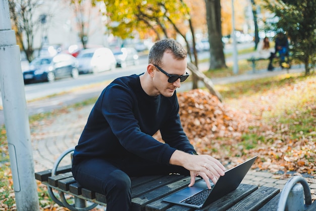 Młody mężczyzna siedzi w parku na ławce i pracuje na laptopie Ciepły jesienny dzień Praca zdalna