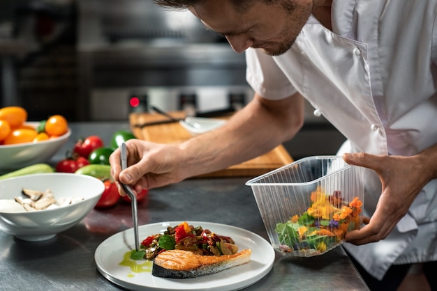 Młody mężczyzna szef kuchni pochyla się nad pieczonymi warzywami na kawałku smażonego łososia na talerzu, dekorując smaczny posiłek przed dostarczeniem do klienta