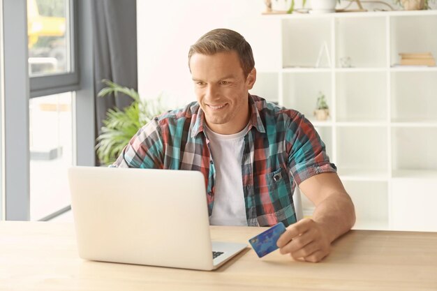 Młody mężczyzna trzyma kartę kredytową podczas korzystania z laptopa w domu