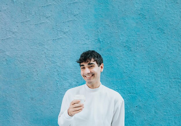 Młody mężczyzna trzymający kawę na wynos nad dużą niebieską ścianą, śmiejąc się do kamery. Koncepcja nowoczesnego stylu życia. Biały, słodszy strój miejski. Dni Madrytu.