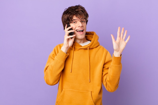 Młody mężczyzna trzymający komórkę uśmiechający się radośnie, machający ręką, witający cię i witający