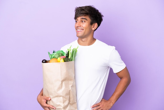 Młody mężczyzna trzymający torbę na zakupy spożywcze na fioletowym tle pozuje z rękami na biodrach i uśmiecha się