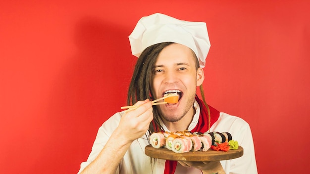 Młody mężczyzna ubrany jak szef kuchni trzymający drewnianą deskę z rolkami sushi na czerwonym tle Mężczyzna kucharz z zamkniętymi oczami je apetyczna rolka sushi pałeczkami