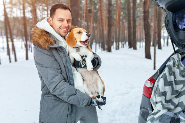 Młody mężczyzna ubrany w szary zimowy park w śnieżnym zimowym lesie trzyma psa o imieniu Beagle.