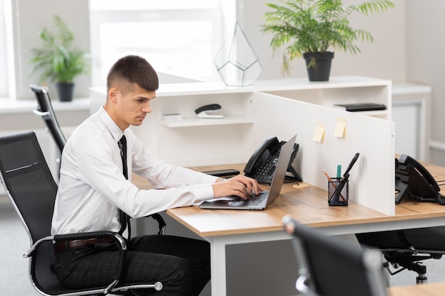 Młody mężczyzna w białej koszuli z czarnym krawatem przy biurku w jasnym biurze pracującym na laptopie