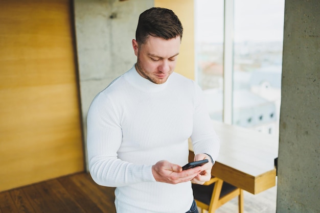 Młody mężczyzna w białym swetrze stoi w przestronnym biurze i rozmawia przez telefon Mężczyzna rozmawia z kolegami za pośrednictwem łącza wideo