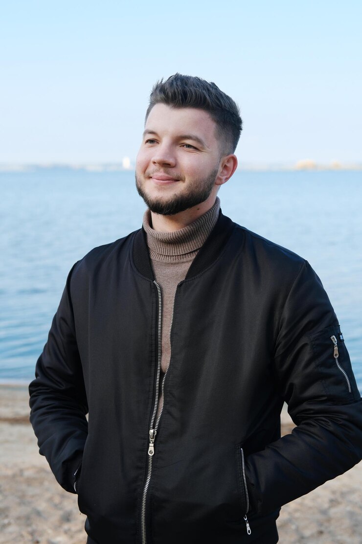 Zdjęcie młody mężczyzna w czarnej kurtce stoi na plaży na tle jeziora i uśmiecha się