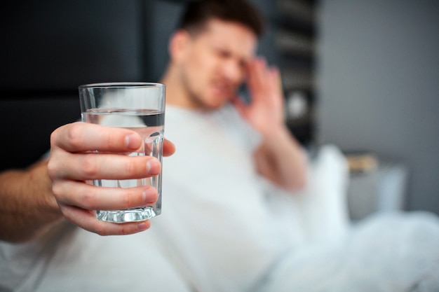 Zdjęcie młody mężczyzna w łóżku trzyma szklankę wody, ma ból głowy lub kaca