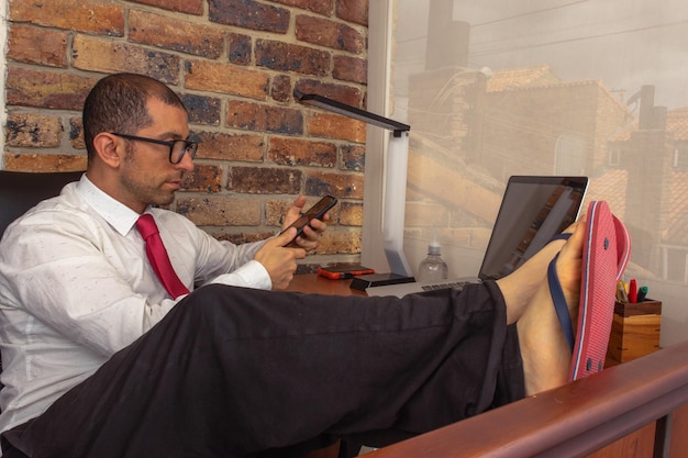 Młody mężczyzna w okularach, krawat i klapki, trzymający smartfona i patrzący na jego laptopa w domowym biurze
