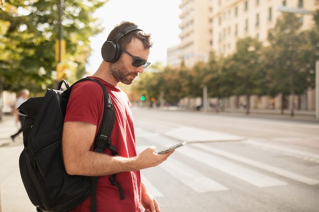 Młody mężczyzna w okularach przeciwsłonecznych, słuchający muzyki i patrzący na swój smartfon podczas wizyty w mieście w Hiszpanii