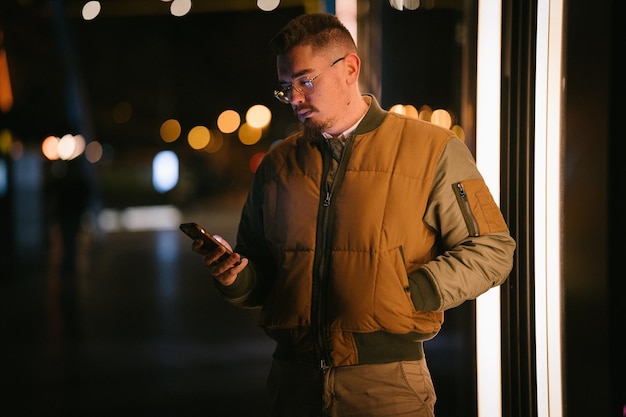 Młody mężczyzna w okularach w nocy, patrząc prosto przed siebie, trzymając telefon w ręku w zimie