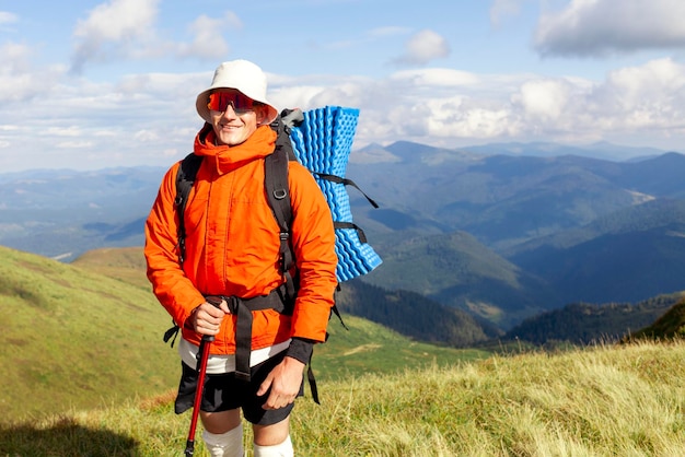 młody mężczyzna w pomarańczowej kurtce i okularach stoi na szczycie góry z plecakiem