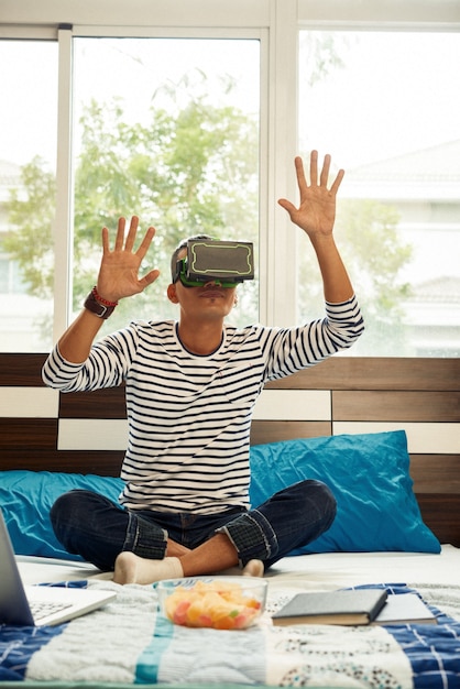 Młody mężczyzna w zestawie wirtualnej rzeczywistości siedzi na łóżku i gra w grę wideo po zakończeniu odrabiania lekcji