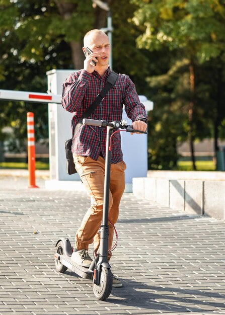 Zdjęcie młody mężczyzna w zwykłych spodniach i koszuli stojący obok swojej elektrycznej hulajnogi i rozmawiający przez telefon komórkowy