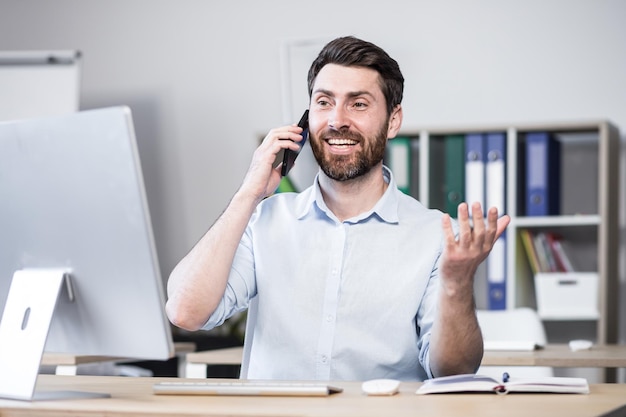 Młody mężczyzna z brodą, pracownik menedżera freelancer rozmawia przez telefon komórkowy w biurze, siedząc przy biurku komputerowym, rozmawiając, machając