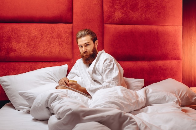 Młody mężczyzna z długimi wąsami i brodą, ubrany w szlafrok, siedzący w łóżku i trzymający krem w rękach. Higieny osobistej.