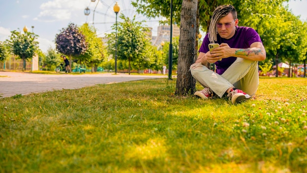 Młody mężczyzna z dredami odpoczywa w cieniu drzewa i używa smartfona w parku Przystojny facet w okularach przeciwsłonecznych siedzi na trawie przy drzewie w słoneczny dzień