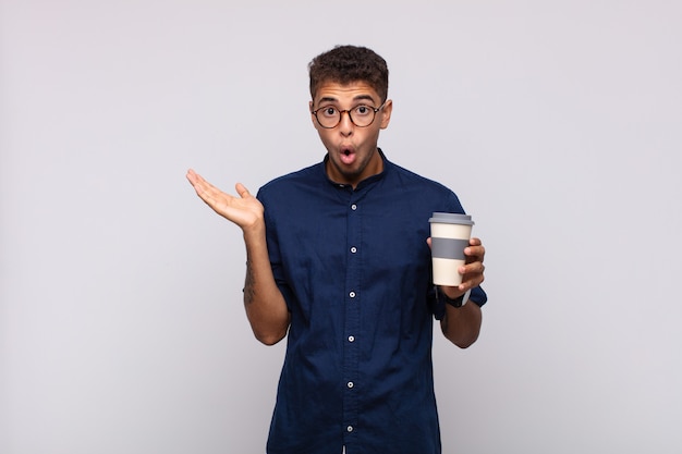 Młody mężczyzna z kawą wyglądający na zaskoczonego i zszokowanego, z opadniętą szczęką, trzymając przedmiot z otwartą ręką na boku